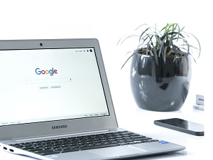 open laptop at the google home screen sitting on a white desk with a plant and a cellphone on desk